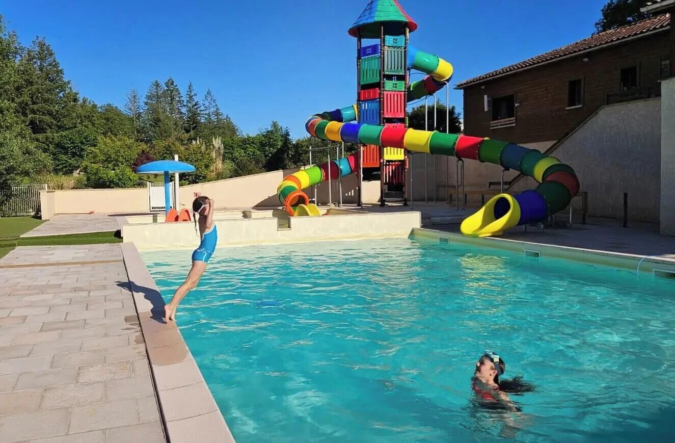 A swimming pool with a slide, a water tower, and a brick building in the background.