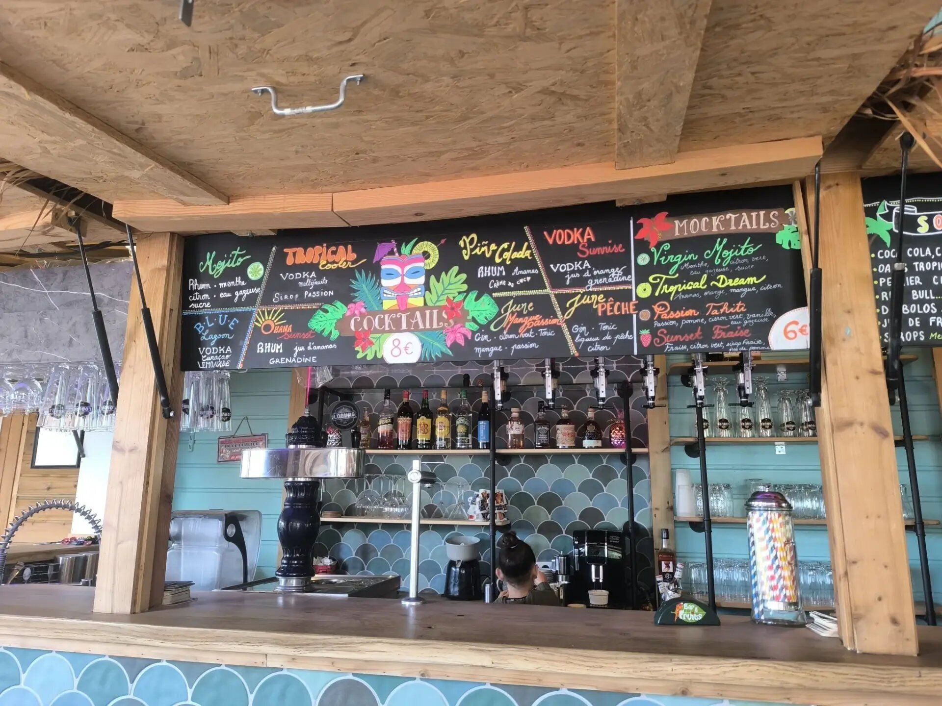 Eine Barbereich mit einer Kreidetafel, auf der Cocktails und Getränke aufgeführt sind, Regale mit Flaschen und Gläsern sowie eine Kaffeemaschine.