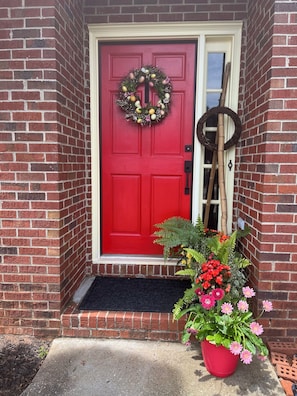 A red door with a wreath, a wooden ladder, and a potted plant with flowers.