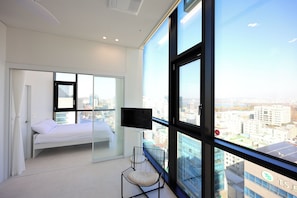 A modern bedroom with a bed, a chair, a TV, and a large window with a city view.