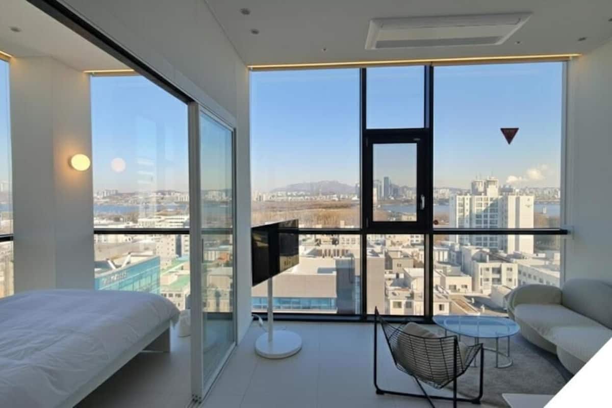 Habitación de hotel en un edificio alto, con un ventanal grande, una cama, una mesita y una silla.