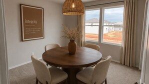Ein Essbereich mit einem runden Tisch, vier Stühlen, einer Hängeleuchte, einer Vase mit Trockenpflanzen, einem gerahmten Zitat an der Wand und Blick durch das Fenster.