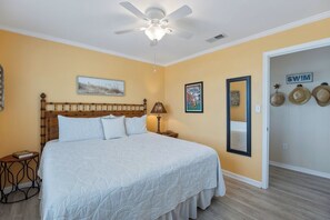 Une chambre avec un lit, un ventilateur de plafond, un tableau encadré, une lampe et une porte sur laquelle est affiché le mot « PISCINE ».