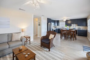 Un salon moderne avec un canapé, une table basse et un fauteuil en osier. On aperçoit à l’arrière-plan une cuisine avec des meubles sombres et un coin repas avec des chaises en bois.