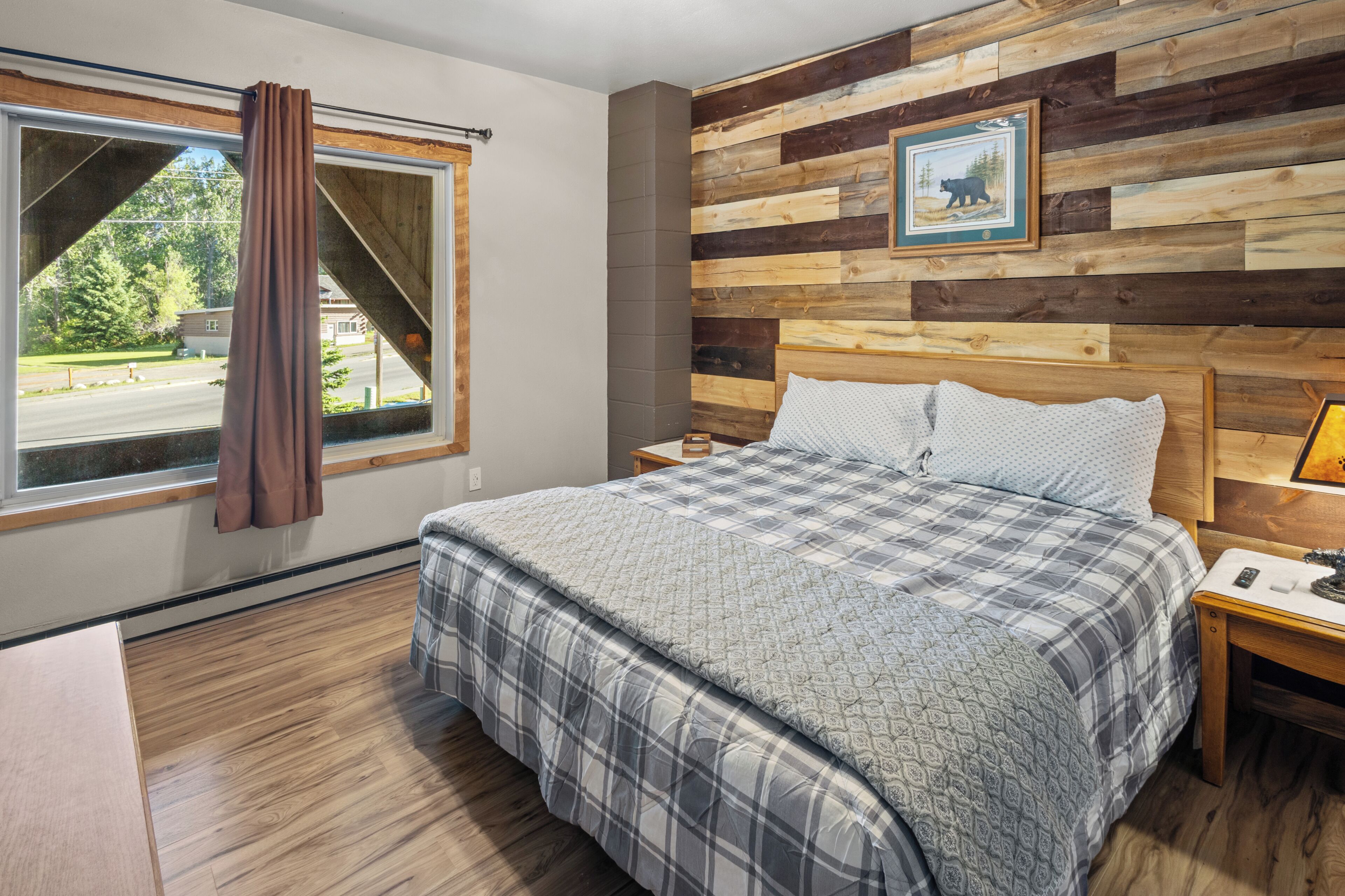 Een slaapkamer met een groot bed, een houten hoofdeinde, een raam met gordijnen, een nachtkastje en een foto aan de muur.