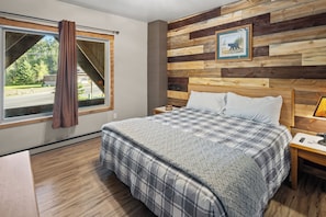 Un dormitorio con una cama grande, cabecera de madera, ventana con cortinas, mesita de noche y un cuadro en la pared.