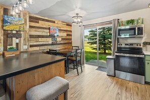 A modern kitchen with a wooden wall, a dining area, and a large window with a view of greenery. - Château Rouge ~ 210 Sawmill Flats (Red Lodge)