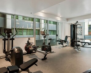A modern gym with various exercise equipment, large windows, and a view of the cityscape. - Free Car Space + Collins 2BR Full Apartment (West Melbourne)