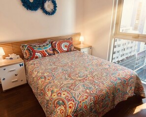 A bedroom with a patterned bedspread, a wooden headboard, two bedside tables with lamps, and a view of buildings through a window. - Free Car Space + Collins 2BR Full Apartment (West Melbourne)
