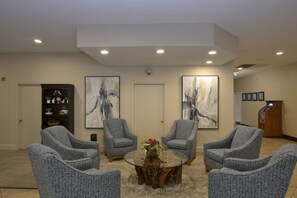 A conference room with four grey upholstered chairs around a glass coffee table, two abstract paintings on the walls, and a podium with a microphone.