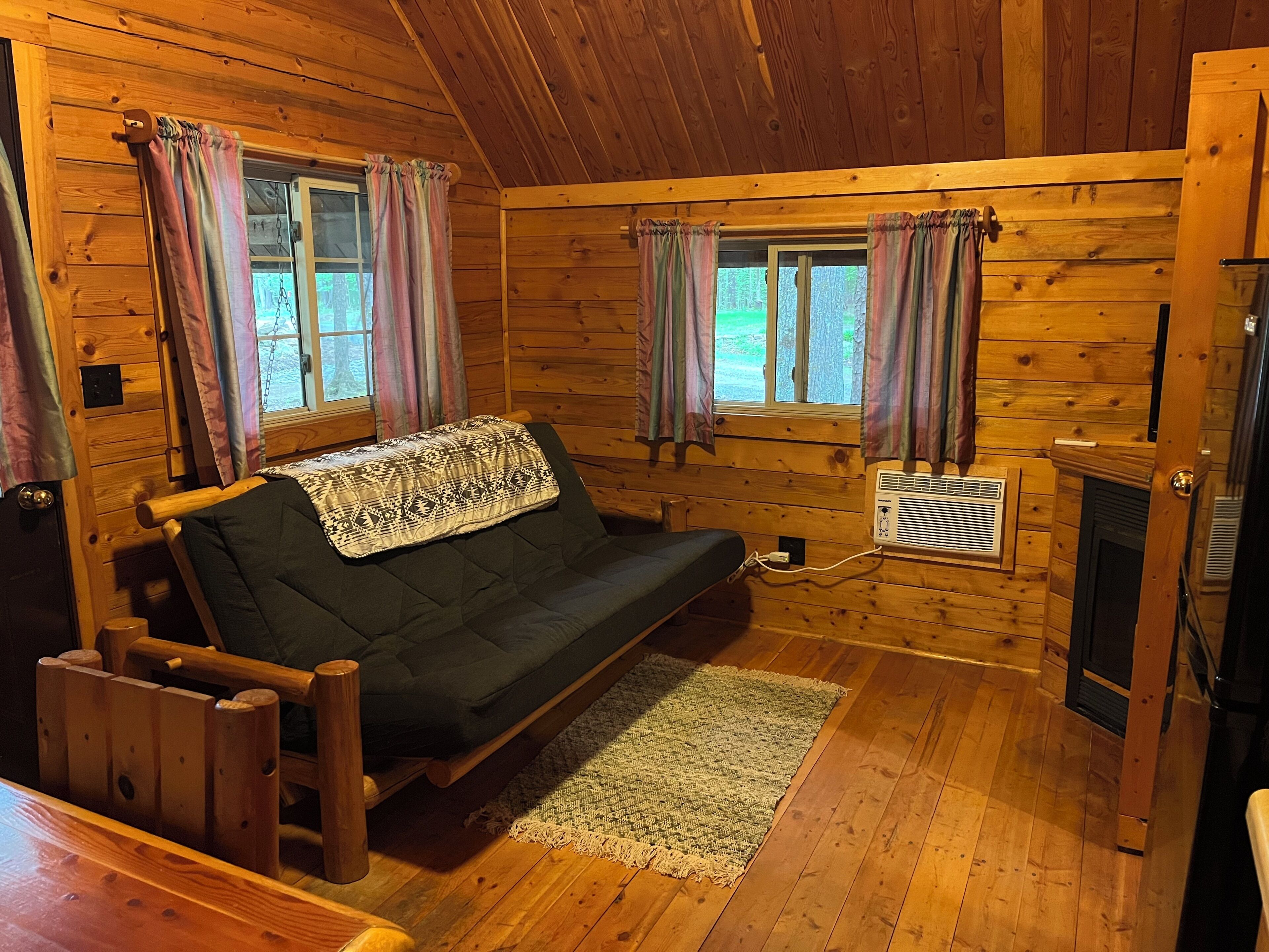 L’intérieur d’un chalet en bois, avec un canapé, deux fenêtres avec des rideaux, une cheminée et un plancher en bois.