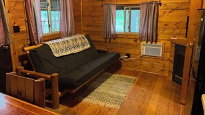 A wooden cabin interior with a sofa, two windows with curtains, a fireplace, and a wooden floor.