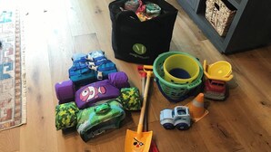 Huoneessa on puulattia, musta logolla varustettu laukku, vihreä ja keltainen kori, leikkukuorma-auto sekä useita värikkäitä koristetyynyjä.
