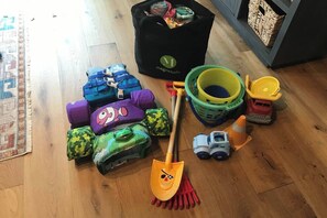 Ein Zimmer mit Holzboden, einer schwarzen Tasche mit Logo, einem grün-gelben Korb, einem Spielzeug-Lkw und mehreren bunten Kissen.