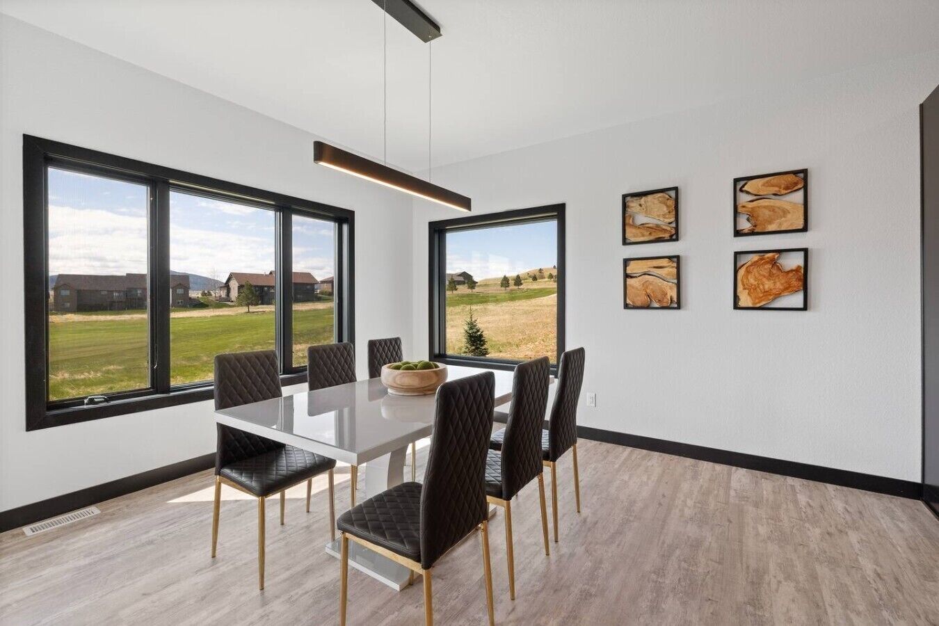 A modern dining room with a large table and chairs, large windows with a view of a grassy field and buildings, and three framed artworks on the wall.
