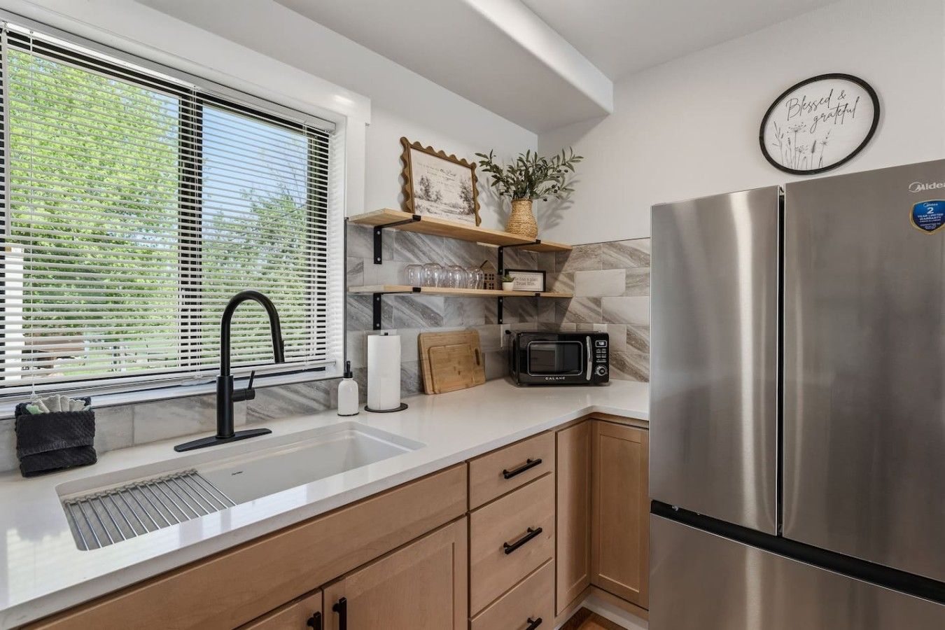 A modern kitchen with a stainless steel refrigerator, a microwave, a sink with a faucet, and a window with blinds.