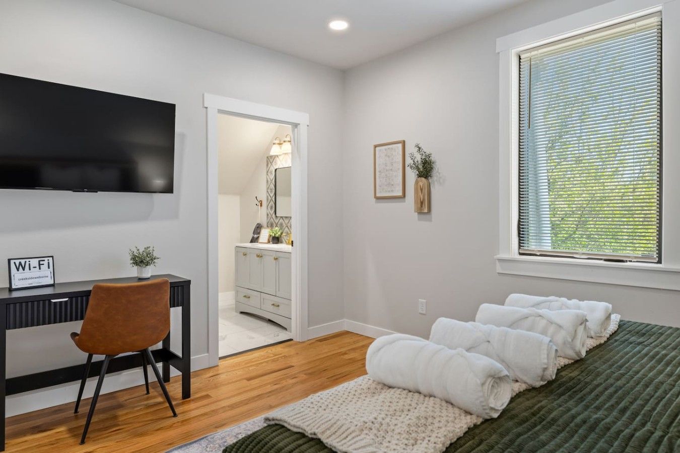 A bedroom with a bed, a desk, a chair, a TV, and a window.