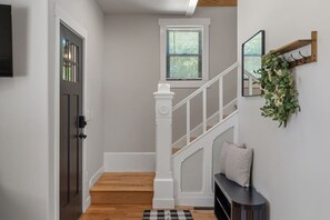 Un couloir avec un plancher en bois, une porte blanche, un escalier avec des rampes blanches, une petite fenêtre avec des stores et un miroir fixé au mur.