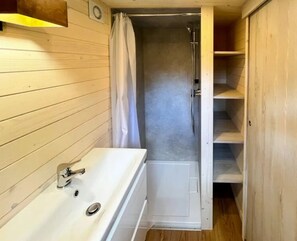 A bathroom with a wooden vanity, a shower, and a door leading to a storage area.