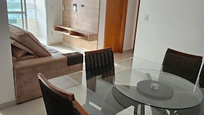 A modern living room with a glass dining table, two chairs, a sofa, and a TV unit. - Family Apartment with 2 Bedrooms in the City Center, 200m from the Beach, Furnished and with AC (Praia Grande)