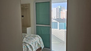 A bedroom with a bed, a sliding door to a balcony, and a view of buildings. - Family Apartment with 2 Bedrooms in the City Center, 200m from the Beach, Furnished and with AC (Praia Grande)