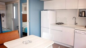 Eine kompakte Küche mit Kühlschrank, Herd, Spüle und Geschirrspüler. Im Vordergrund ist ein Esstisch mit einem Stuhl zu sehen.