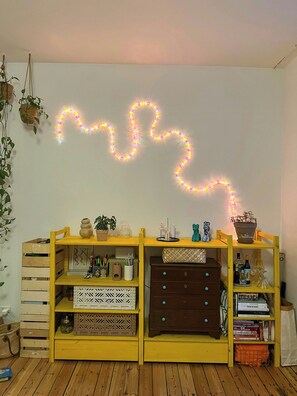 Ein Zimmer mit Holzboden, einer gelben Regaleinheit mit verschiedenen Gegenständen und einer Lichterkette, die eine Herzform an der Wand bildet.