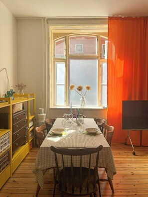 Ein Essbereich mit einem Tisch für vier Personen, einem Fenster mit Blick auf ein Ziegelgebäude und einem Fernseher auf einem Schrank.