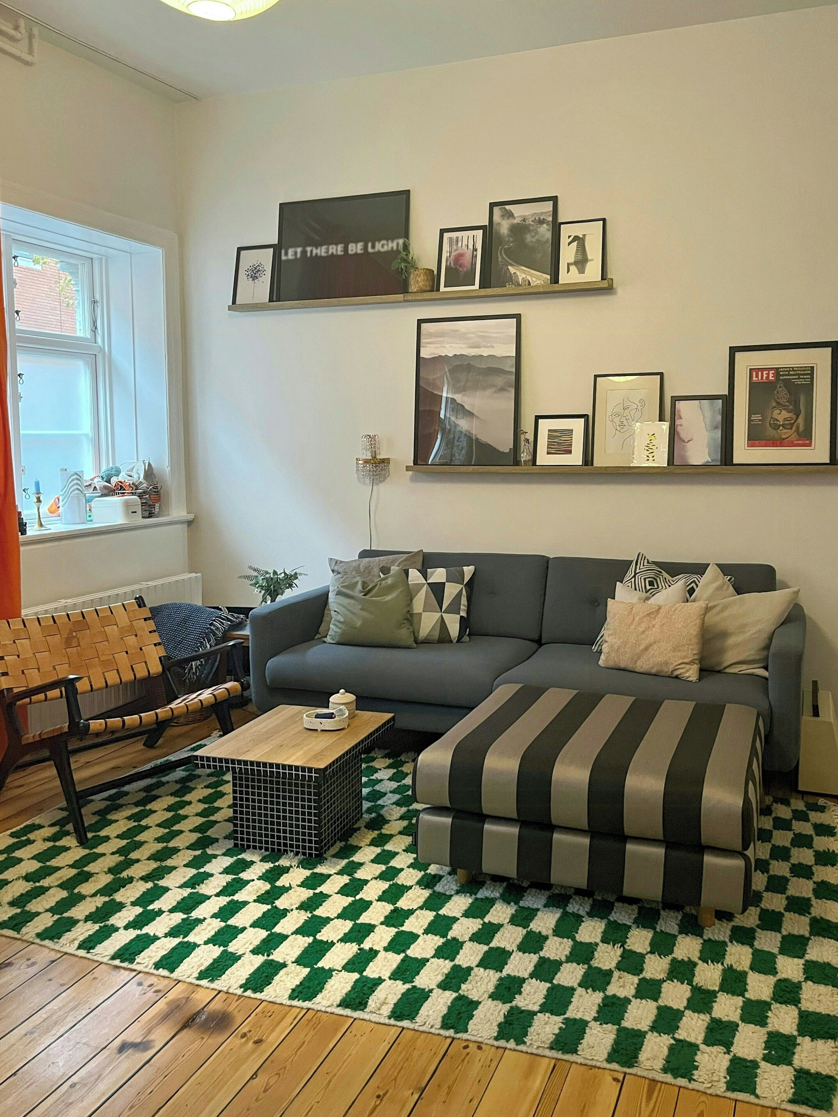 Ein Wohnzimmer mit einer Couch, einem gestreiften Ottomanen, einem Holz-Couchtisch und einem karierten Teppich. An der Wand befinden sich gerahmte Bilder und ein Fenster mit Blick.