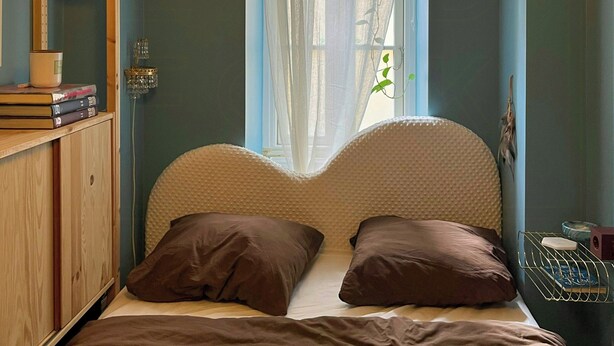 Ein Schlafzimmer mit einem Bett, einem Holzschrank, einer Pendelleuchte und einem Fenster mit transparenten Vorhängen.
