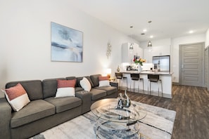 A modern living room with a grey sofa, a glass coffee table, and a kitchen area with white cabinets and a bar. - Cozy 1-bedroom apartment near FIFA, universal studio resort in tranquil Frisco (Frisco)