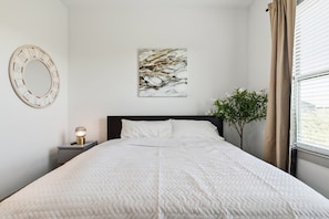 A neatly made bed with a quilted white duvet, a wall-mounted round mirror, a painting, a bedside lamp, and a potted plant. - Cozy 1-bedroom apartment near FIFA, universal studio resort in tranquil Frisco (Frisco)