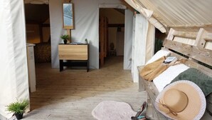 Un alojamiento con carpa, suelo de madera, una cama con manta, un sombrero, pantuflas sobre un banco y una planta pequeña en el suelo.