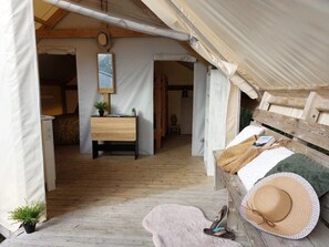 A tented accommodation with a wooden floor, a bed with a blanket, a hat, and slippers on a bench, and a small plant on the floor. - Camping 3 étoiles - Piscine  - ccbfidh (Les Moutiers-en-Retz)