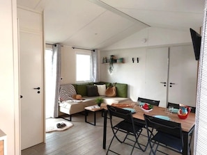 A cozy living room with a sofa, a dining table with chairs, a small coffee table, and a window with curtains. - Camping 3 étoiles - Piscine  - ccbfie0 (Les Moutiers-en-Retz)