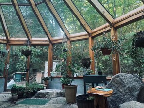 Une serre avec une structure en bois, des plantes en pot, une table avec un carnet et un rocher.