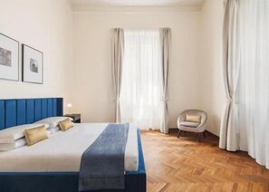Hotellihuone, jossa on suuri sänky, tuoli, yöpöytä ja ikkuna, jossa on verhot.