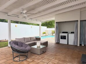 Eine überdachte Terrasse mit Deckenventilator, einem grauen Ecksofa, einem Korbstuhl, einem Tisch mit Glasplatte und einem Pool im Hintergrund.