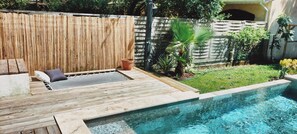 Un espace aménagé au bord de la piscine, avec une terrasse en bois, un coin salon doté de coussins et un petit palmier.