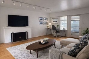 A modern living room with a fireplace, a flat-screen TV, a desk with a chair, and a sofa.