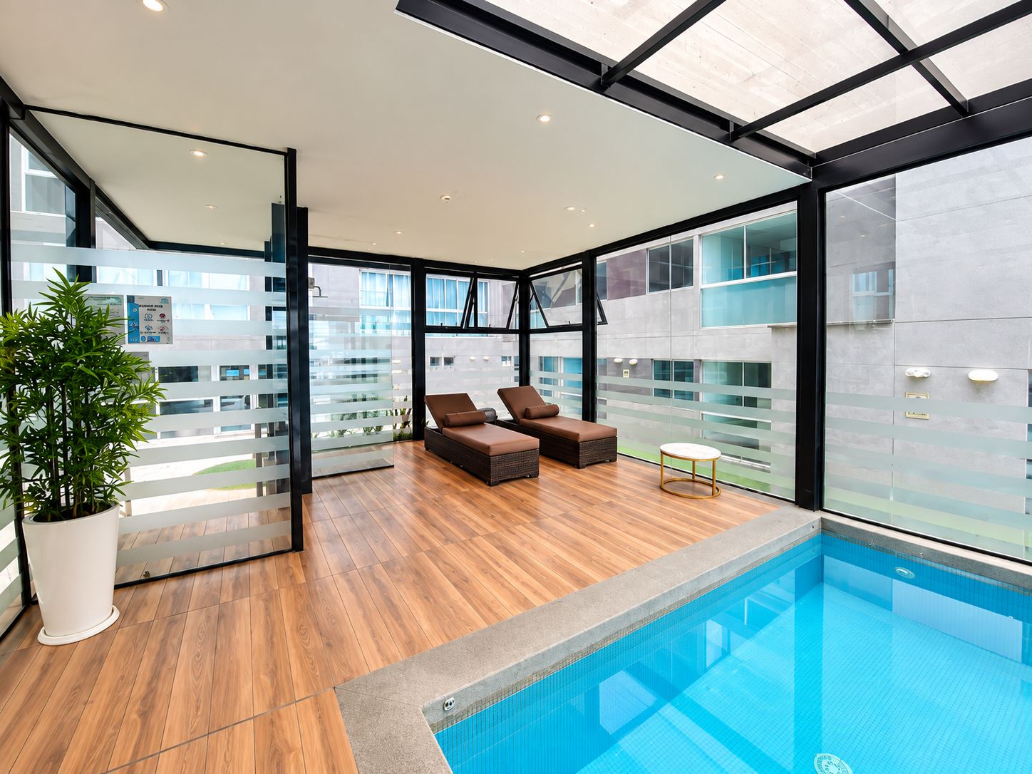 A modern indoor pool area with wooden flooring, two lounge chairs, and a glass-enclosed space with a view of the city.