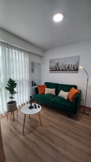 Una sala de estar con un sofá verde, una mesa de centro redonda, una planta en maceta y un cuadro de un paisaje urbano en la pared.