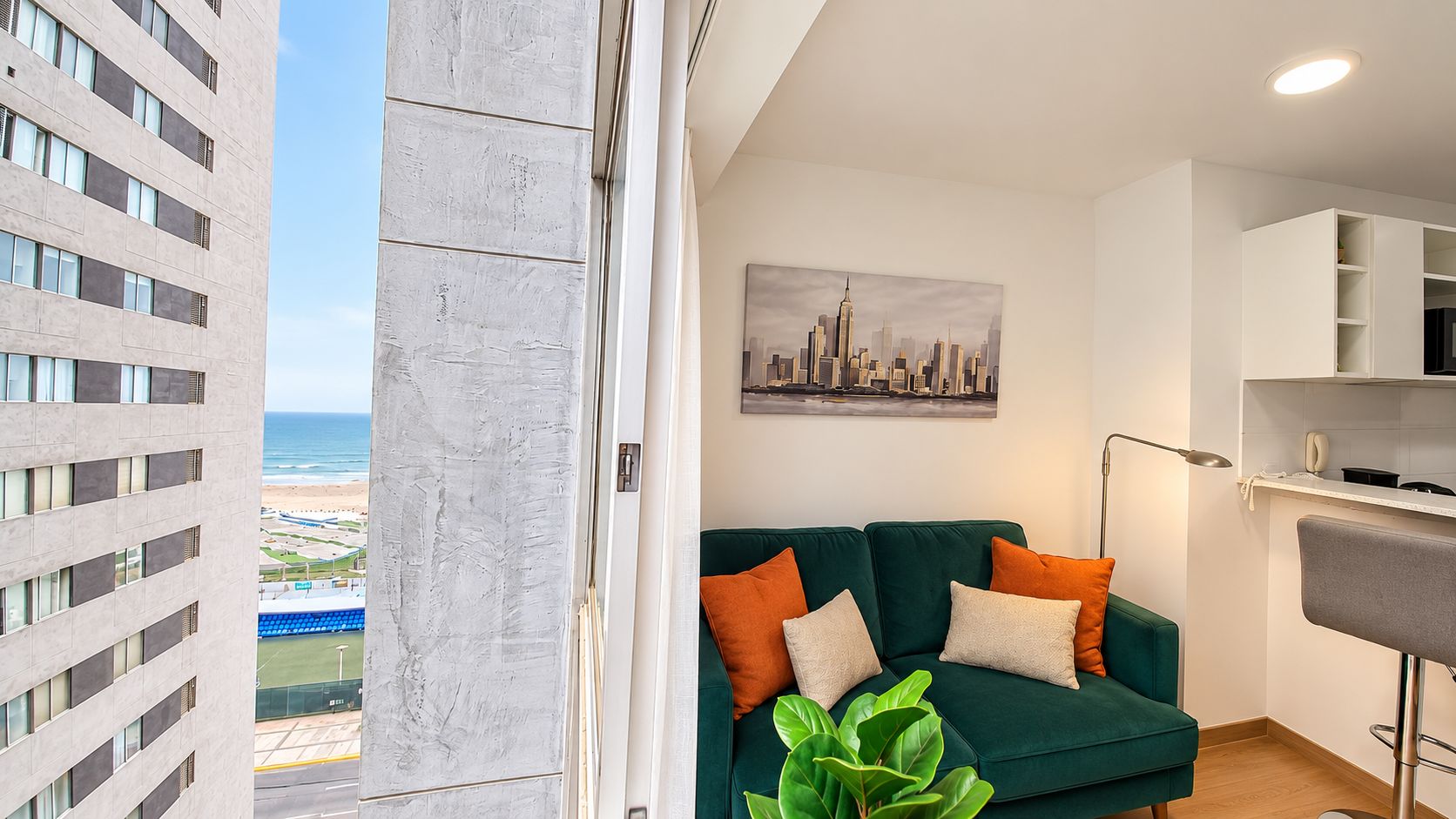 Una sala de estar moderna con un sofá verde, una pintura de un paisaje urbano y vistas a una playa y edificios.