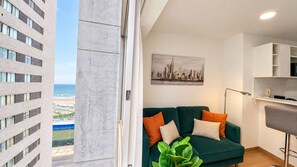 A modern living room with a green sofa, a cityscape painting, and a view of a beach and buildings. - Loto Stays Apartment across from Costa21 | Arena1 | Self-check-in (San Miguel)