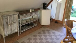 Una habitación rústica con un armario de madera, un refrigerador blanco pequeño, una alfombra a cuadros y un sillón de mimbre.