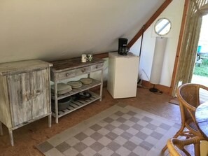 Ein rustikaler Raum mit einem Holzkabinett, einem kleinen weißen Kühlschrank, einem karierten Teppich und einem Korbstuhl.