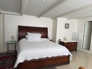 A bedroom with a wooden bed, white bedding, a nightstand with a lamp, a bedside table, and a red patterned rug. - LA JOLLA PRIVATE STUDIO (San Diego)