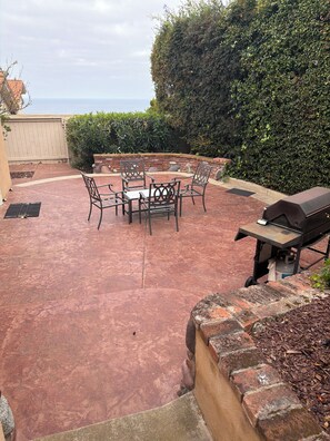 Une terrasse extérieure avec une table ronde et des chaises, un barbecue et une vue sur l’océan.