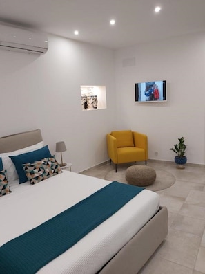 Een moderne slaapkamer met een bed, een gele fauteuil, een klein rond voetenbankje, een tv aan de muur en een kamerplant.