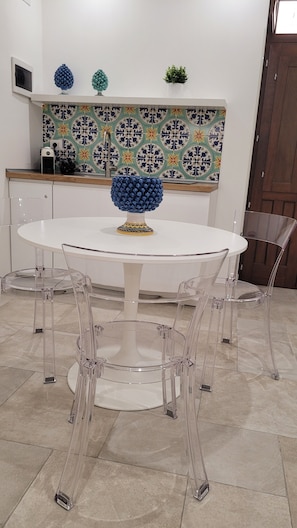 Een ronde tafel met een decoratieve schaal, transparante stoelen, een betegelde achterwand en een houten deur.